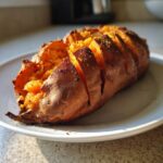 A fluffy baked sweet potato, scored and seasoned, resting on a white plate.