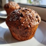 A close-up, sunlit photo of a rich, dark chocolate chip muffin topped with coarse sea salt.