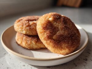 Three soft churro cookies heavily coated in cinnamon sugar resting on a small, light-colored ceramic plate.