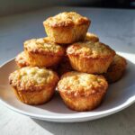 A stack of freshly baked, golden brown mini banana muffins piled high on a white plate.