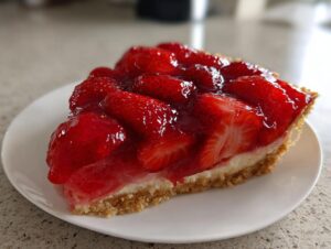 Close-up of a thick slice of strawberry pie featuring a graham cracker crust, cream filling, and a glossy topping of fresh strawberries.
