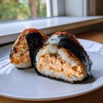 Close-up of two seasoned tuna rice balls (onigiri) wrapped in nori, one cut in half showing the filling.
