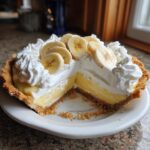 A whole banana cream pie with one slice removed, topped with whipped cream and fresh banana slices.