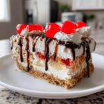 A decadent slice of banana split cake featuring layers of crust, cream, bananas, and topped with whipped cream, chocolate syrup, and maraschino cherries.