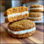 Two carrot cake cookies stacked, one cut in half to show the thick cream cheese frosting filling.