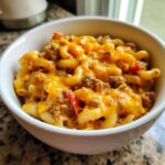 A close-up of a white bowl filled with cheesy taco pasta, featuring elbow macaroni, ground meat, and melted cheddar cheese.