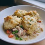 A white bowl filled with creamy chicken and dumplings with biscuits, peas, and carrots, garnished with parsley.