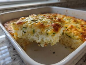 A serving spoon lifts a portion of the golden-brown chicken zucchini casserole from the white baking dish.