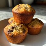 A stack of four golden brown chocolate chip banana muffins sitting on a white plate in bright sunlight.