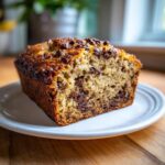 A moist slice of chocolate chip vegan banana bread with a golden brown crust, served on a white plate.