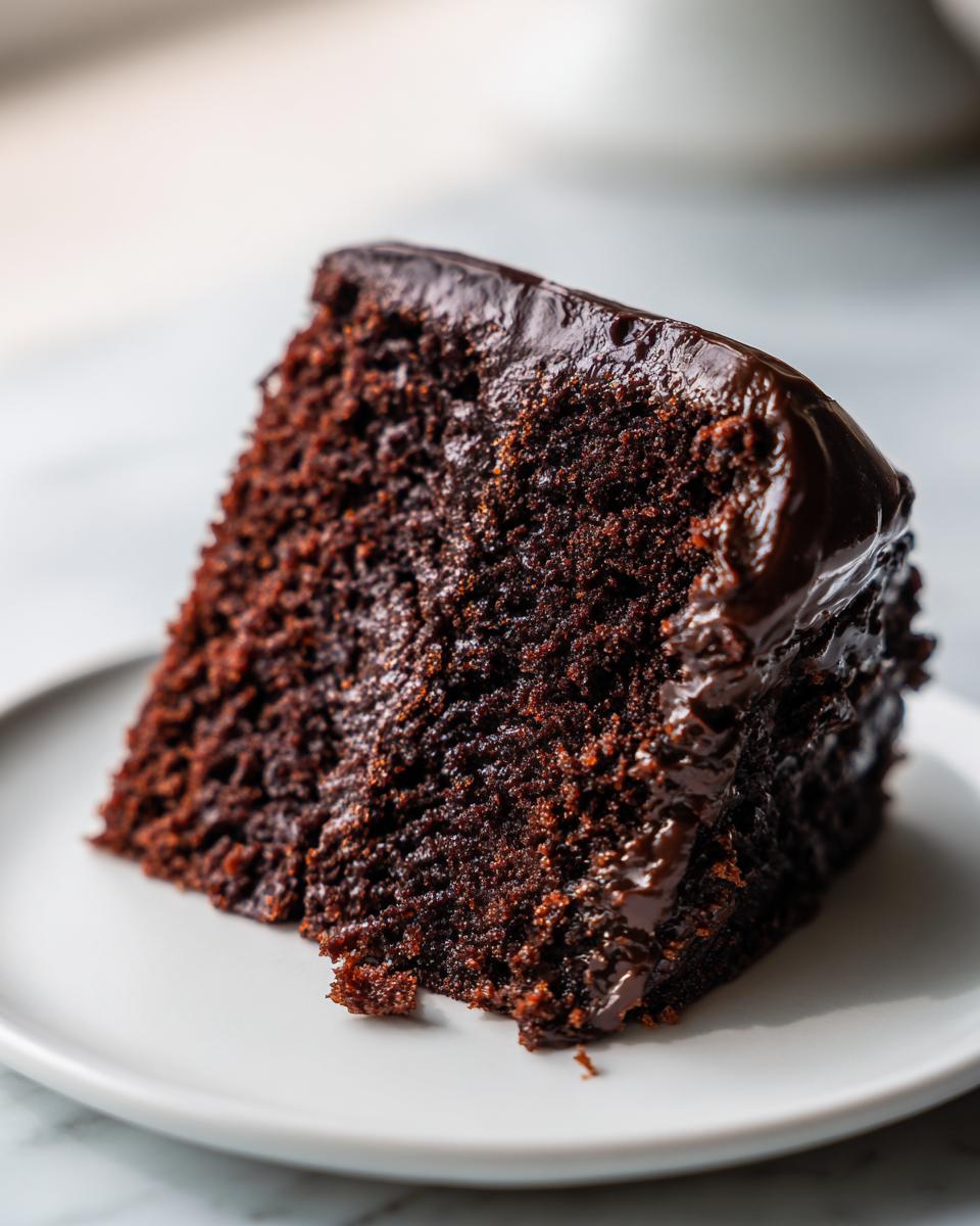 Close-up of a rich, dark slice of chocolate zucchini cake topped with shiny chocolate ganache.