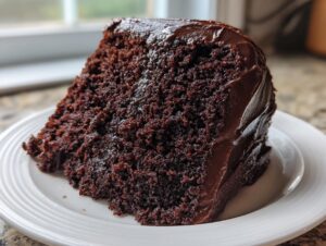 A close-up of a rich, dark slice of chocolate zucchini cake topped with thick chocolate frosting on a white plate.
