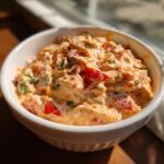 Close-up of creamy, orange-hued boat dip mixed with chunks of red pepper and green onion in a white bowl.