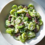 Close-up of a creamy salad featuring sliced green Brussels sprouts, dark raisins, and grated cheese, resembling a celery salad.