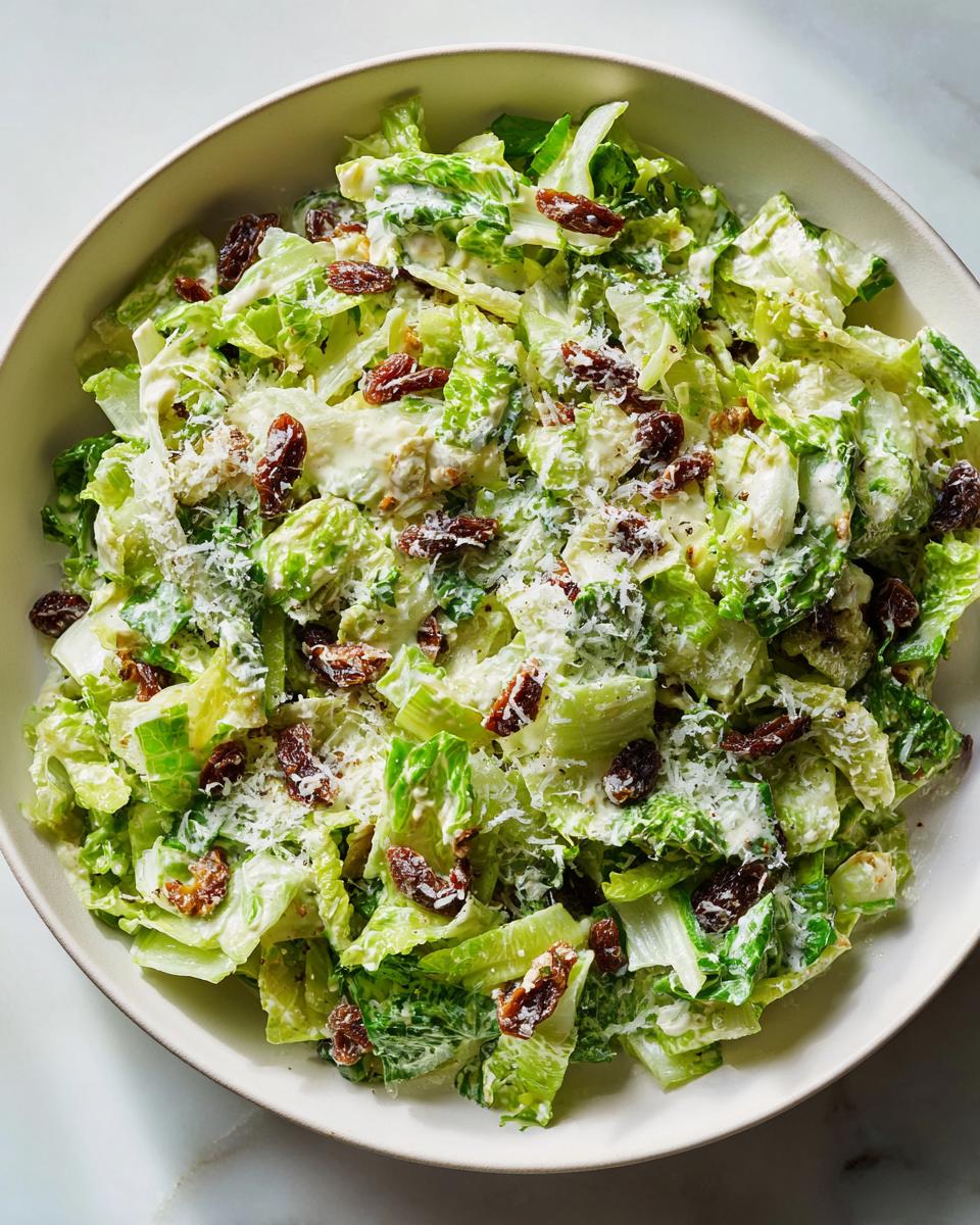 Overhead view of a bowl filled with creamy celery salad, topped with shredded cheese and dark raisins.