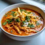Close-up of a bowl of creamy chicken enchilada soup topped with shredded cheese, sour cream, cilantro, and tortilla strips.