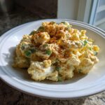 A close-up of creamy potato salad mixed with green onions and topped with a dusting of paprika.