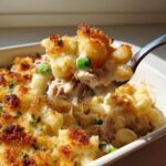 A spoonful of creamy tuna pasta with peas and a crispy, cheesy topping being lifted from a baking dish.