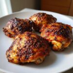 Four perfectly cooked, juicy chicken thighs with dark, crispy, seasoned skin resting on a white plate.