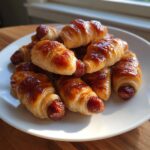 A pile of shiny, glazed pigs in a blanket appetizers served on a white plate.