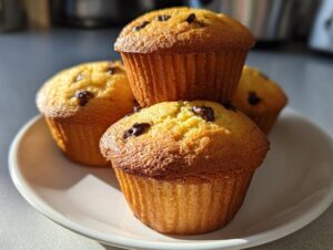 Four golden brown chocolate chip banana muffins stacked slightly on a white plate in bright sunlight.