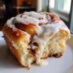 A close-up of a soft, freshly baked cinnamon roll with thick white icing dripping down the side.