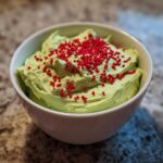 Close-up of bright green Grinch dip in a white bowl, topped generously with small red sprinkles.
