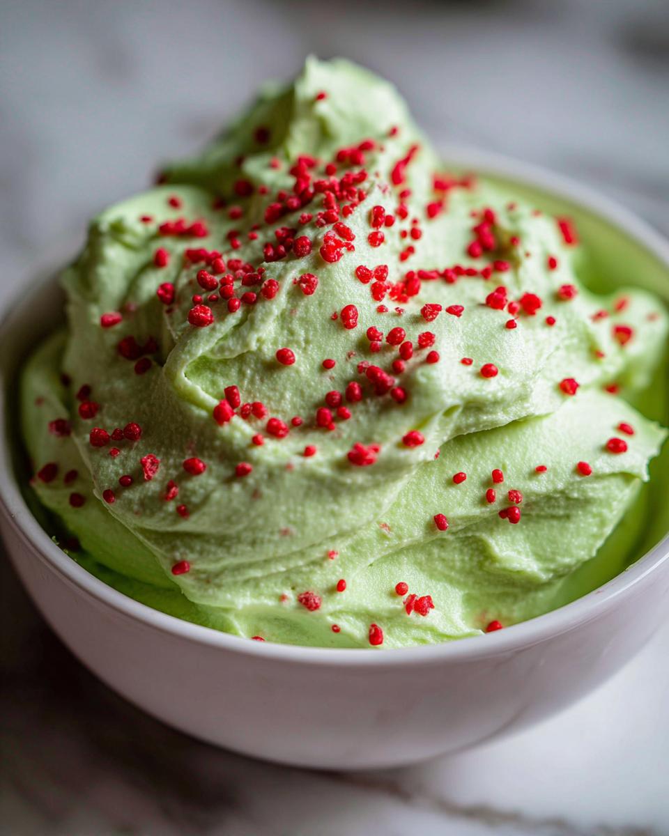 A close-up of bright green grinch dip swirled high in a white bowl and topped with small red sprinkles.