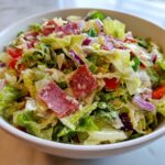 A close-up of a vibrant grinder salad in a white bowl, featuring chopped lettuce, salami, red onion, and creamy dressing.