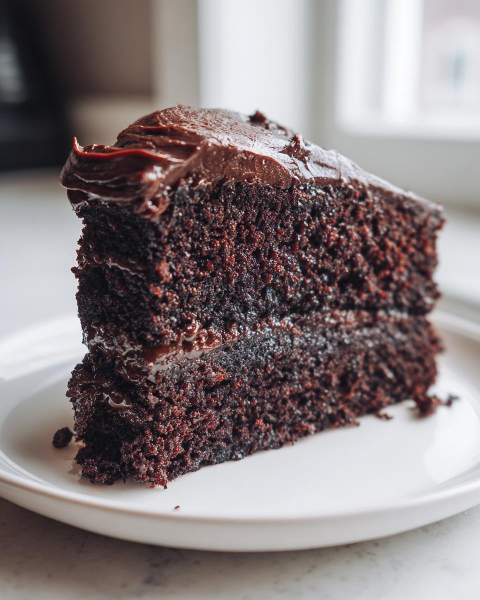 A thick, moist slice of dark chocolate zucchini cake layered with rich chocolate frosting on a white plate.
