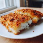 Two pieces of flaky, white fish covered in a golden brown parmesan crusted tilapia topping on a white plate.