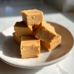 A stack of creamy, light brown squares from a peanut butter fudge recipe sitting on a speckled white plate.
