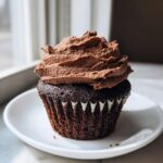 A single, moist chocolate cupcakes topped with a generous swirl of rich chocolate frosting, sitting on a white plate.