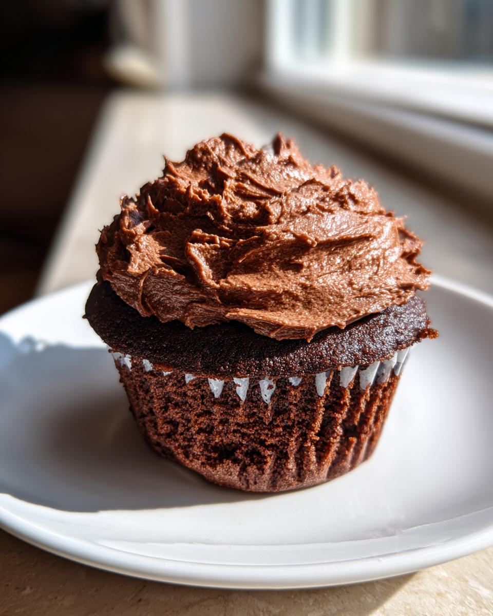 A single, dark chocolate cupcake topped with a generous swirl of rich chocolate frosting, sitting on a white plate.