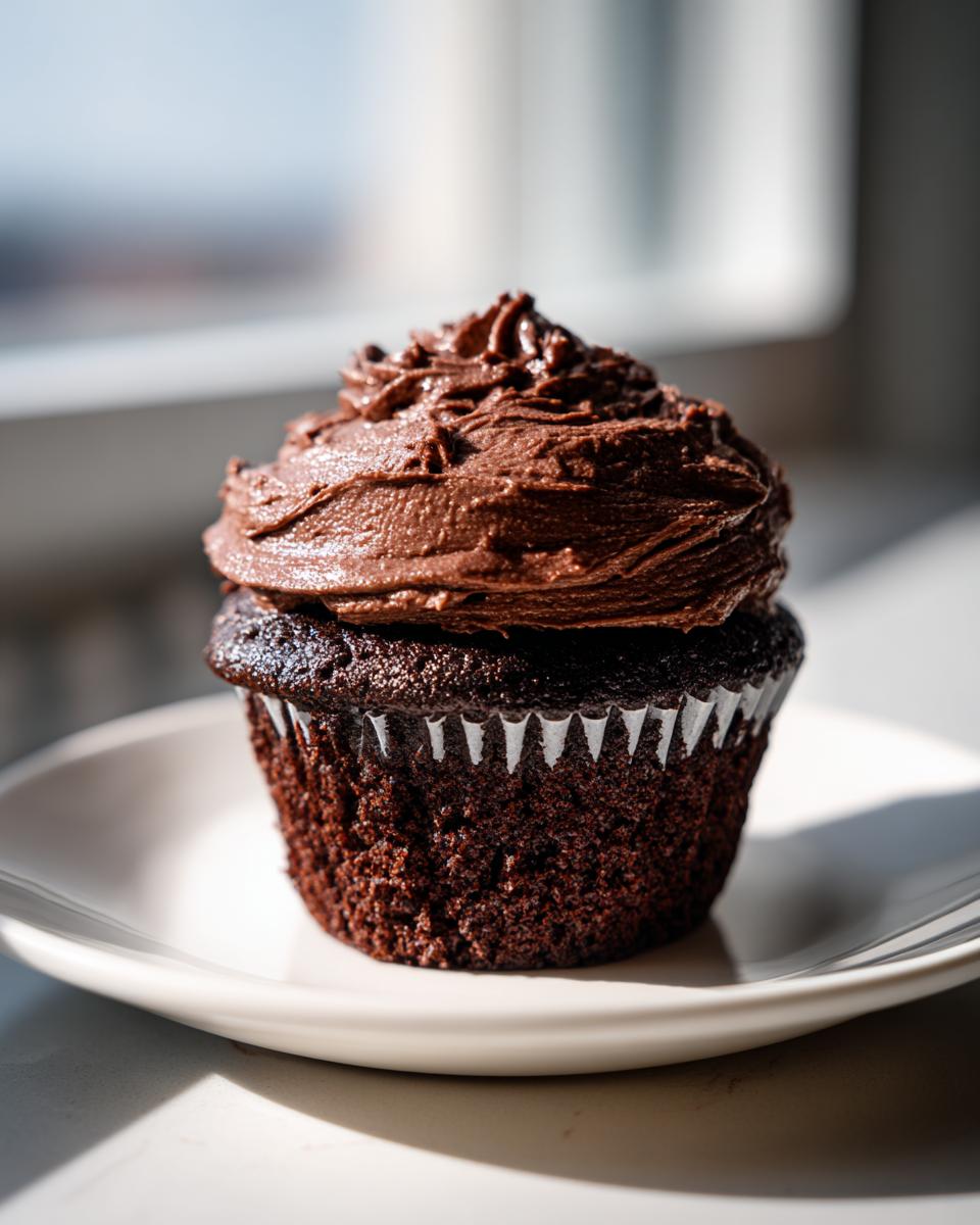 A single, dark chocolate cupcake topped with a generous swirl of rich chocolate frosting, sitting on a white plate.