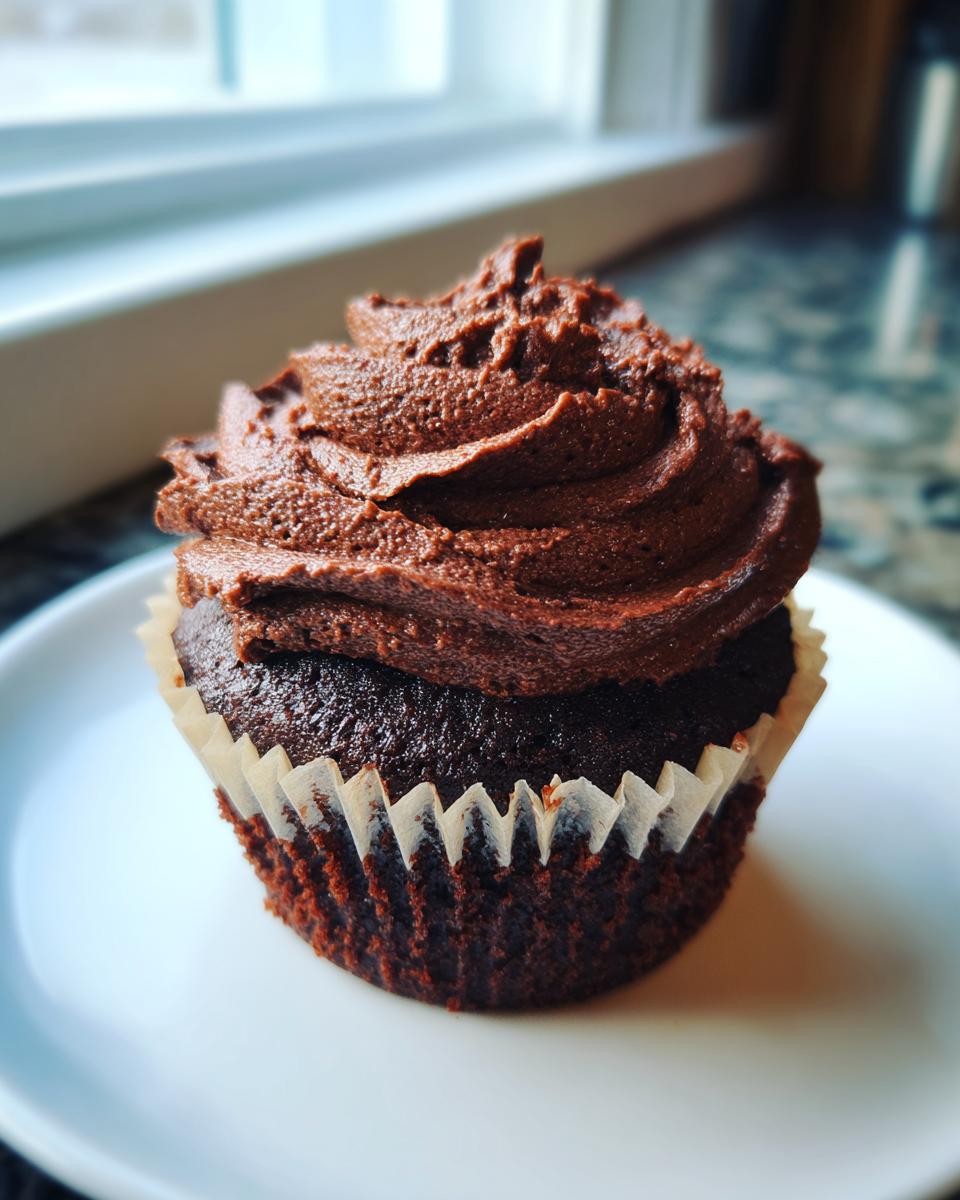 A single, moist chocolate cupcake topped with thick, dark chocolate frosting, sitting on a white plate.