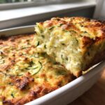 A square slice of baked chicken zucchini casserole being lifted from a white baking dish, showing a cheesy, creamy interior.