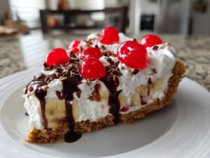 A decadent slice of banana split cake featuring a graham cracker crust, bananas, whipped topping, chocolate syrup, and maraschino cherries.