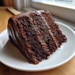 A thick slice of moist, dark chocolate cake recipe layered with rich chocolate frosting on a white plate.