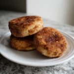 Three soft churro cookies stacked on a white plate, generously coated in cinnamon sugar.