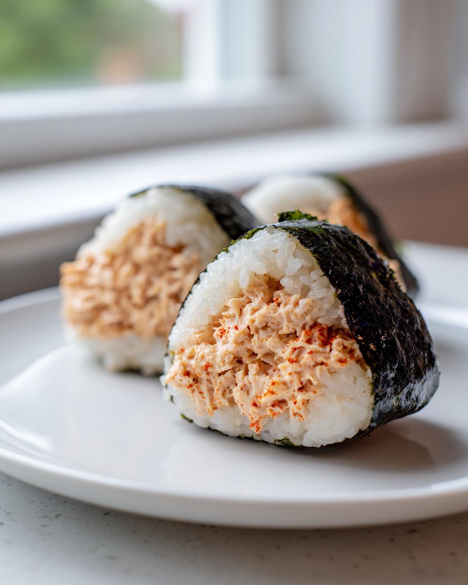 Three triangular tuna rice balls (onigiri) filled with seasoned tuna salad, wrapped partially in dark nori seaweed, served on a white plate.