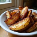 Close-up of golden brown, crispy seasoned potato wedges piled high in a white serving bowl.