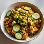 A bowl of seasoned crispy rice salad mixed with shredded chicken, topped with diced avocado and fresh cucumber slices.
