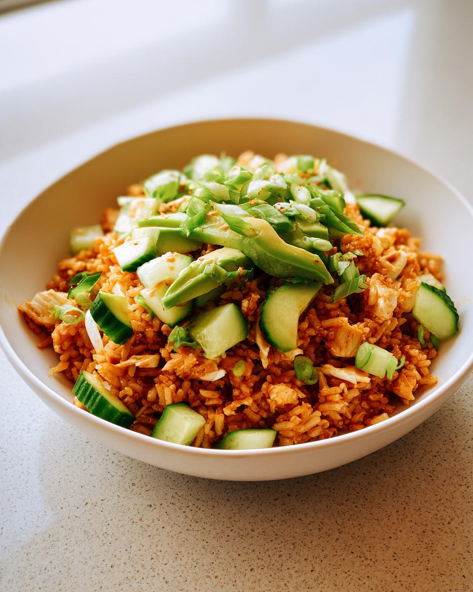 A close-up of a white bowl filled with vibrant orange crispy rice salad, topped with sliced avocado, cucumber chunks, and green onions.