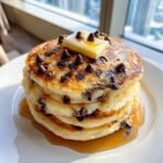 A tall stack of three fluffy chocolate chip pancakes topped with melting butter and syrup.