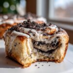 A close-up shot of a delicious oreo cinnamon rolls with a bite taken out, covered in white icing and dark cookie crumbles.