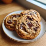 Three thick, chewy chocolate chip cookie rounds topped with flaky sea salt on a light plate.