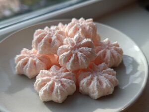 A pile of light pink, star-shaped cream cheese mints dusted with powdered sugar, sitting on a white plate near a window.