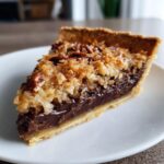 A close-up of a rich slice of german chocolate pie featuring a dark chocolate layer and a toasted coconut pecan topping.
