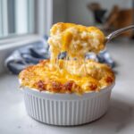 A spoonful of hot, creamy cheesy rice is lifted from a white ramekin, showing long, gooey cheese pulls.